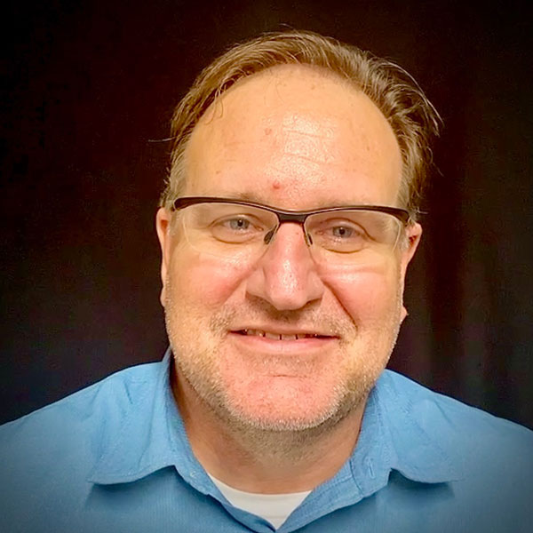An image of Tim Bluhm with a dark background. Tim is wearing a blue shirt and wearing glasses in this picture.
