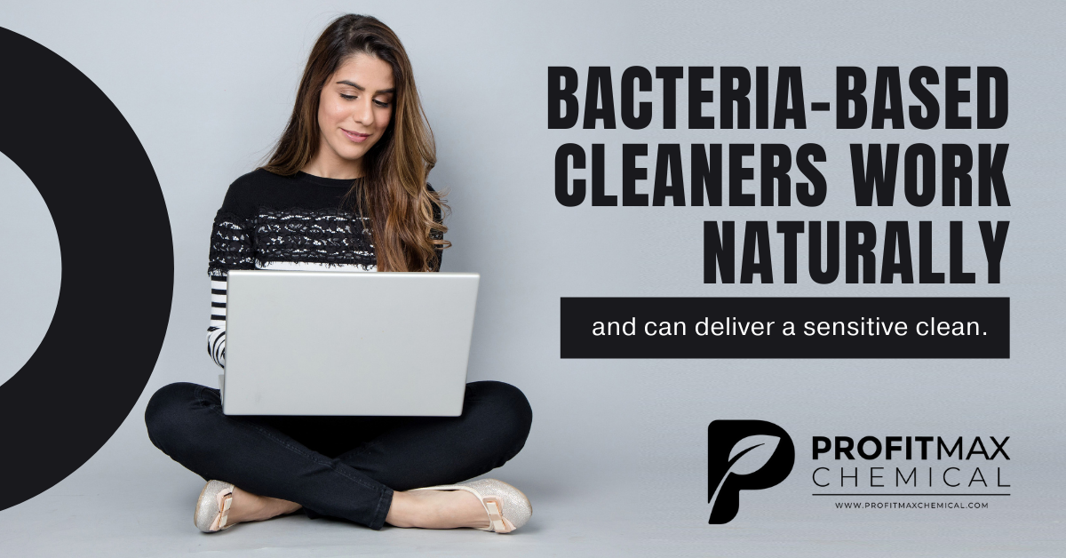 A gray rectangular background with a half shaped oval on the left side. In the middle is a woman at her large lap top smiling with long brown hair and a sweater, pants, and ballet type shoes. To the right of her in black text, it reads: Bacteria-based cleaners can work naturally. Beneath that in a black box is white text that reads: and can deliver a sensitive clean. In the lower right corner is the ProfitMax Chemical Logo P in black with the text ProfitMax Chemical and the website under a line in black text that reads: www.profitmaxchemical.com.